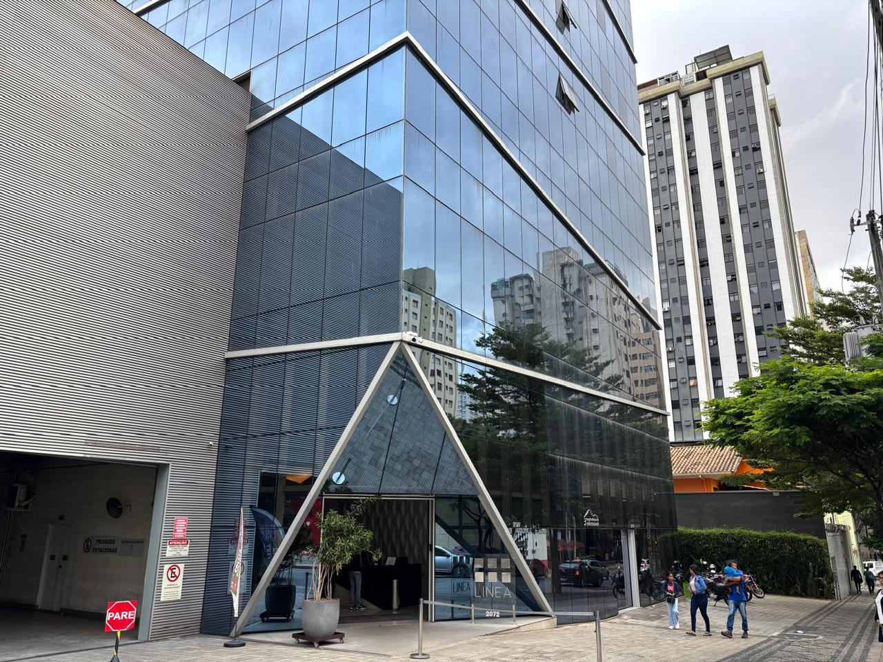 Prédio de escritórios moderno com fachada de vidro espelhado refletindo prédios vizinhos, entrada triangular e pessoas caminhando na calçada.