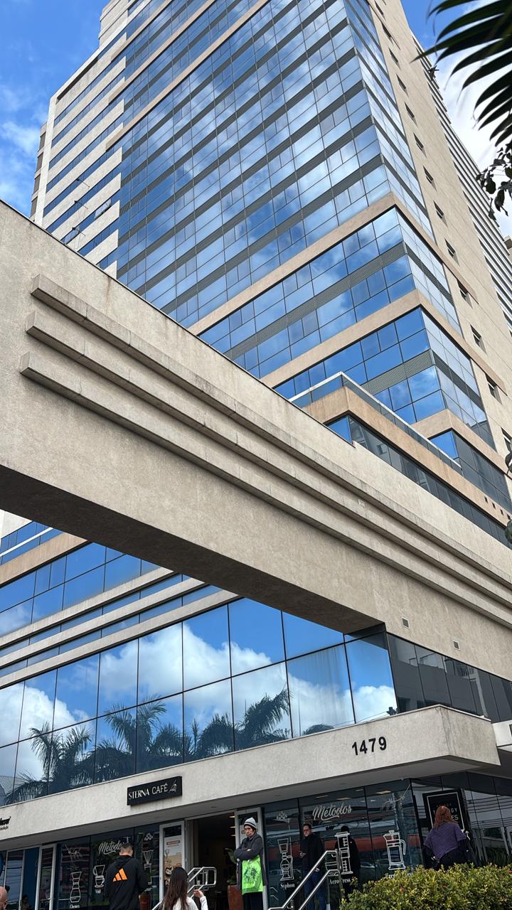 Edifício comercial alto com grandes janelas de vidro refletindo o céu azul e nuvens, com a entrada do Sierna Café e pessoas do lado de fora.