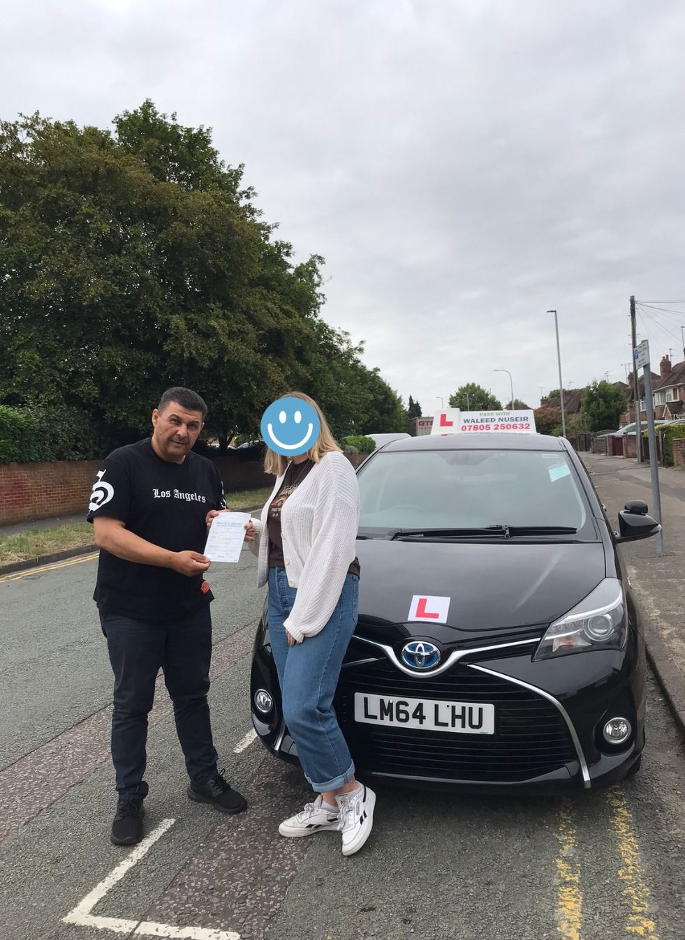 Man and woman standing in front of a black Toyota driving school car on a street, holding a document.