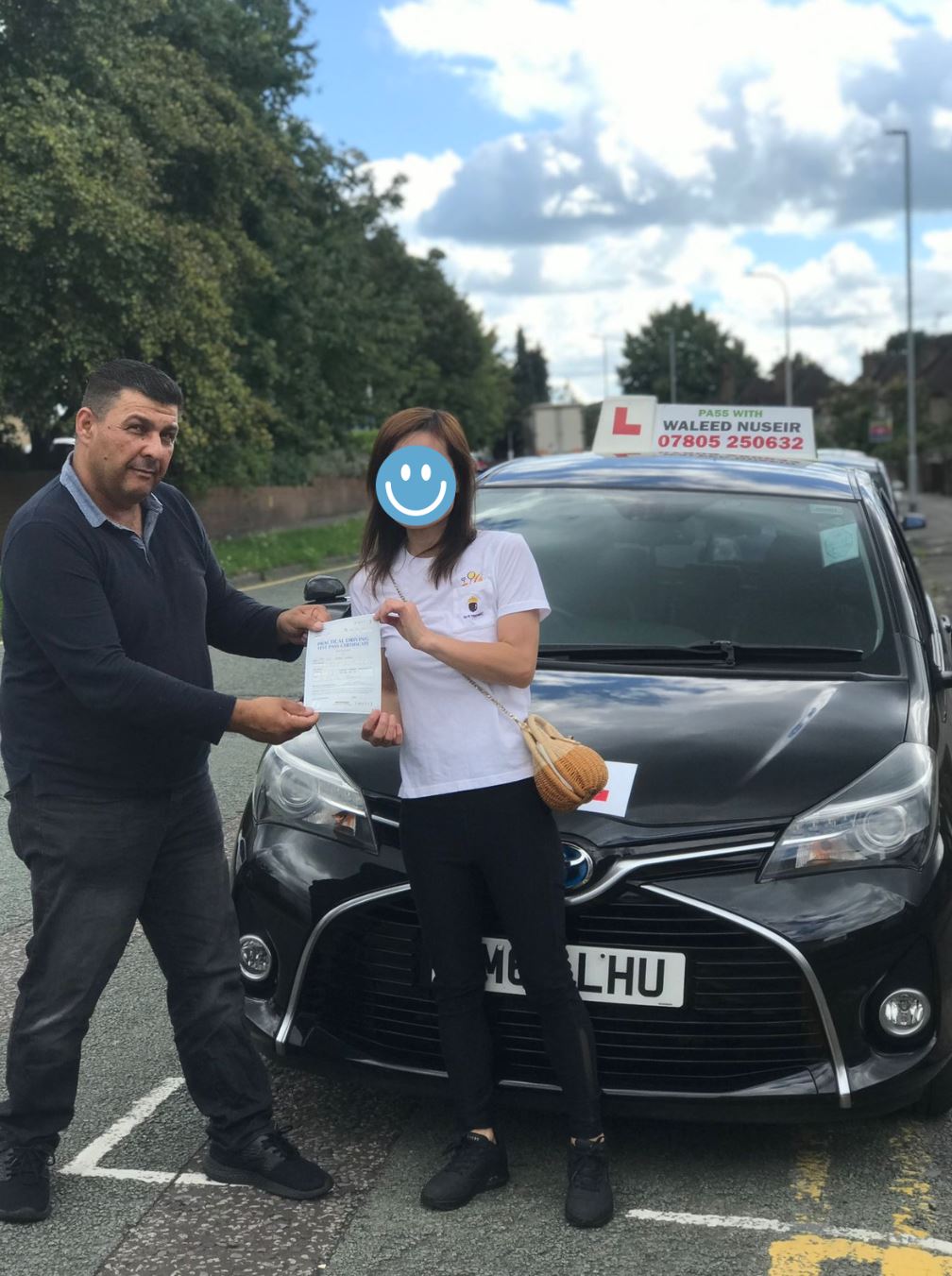 A man and woman standing in front of a black driving school car; the woman holds a certificate, and the car has a learner driver sign on the roof.