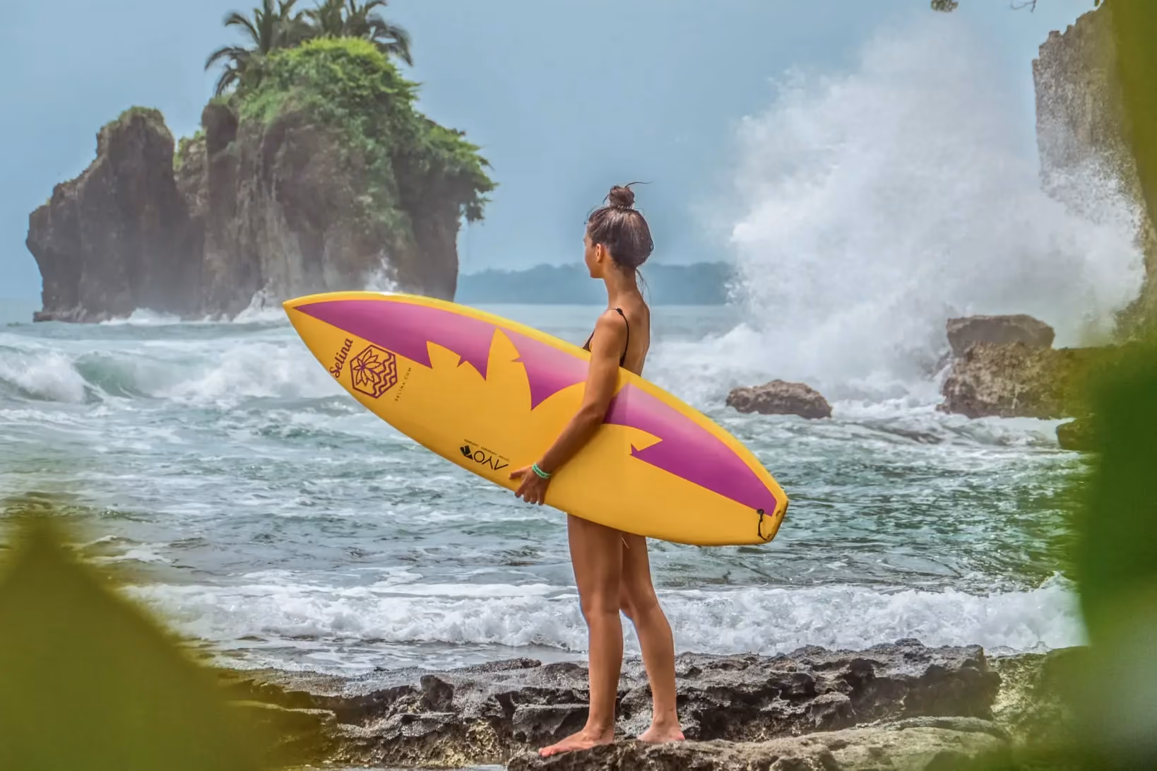 Surfer holding a yellow and purple board standing on rocky shore watching waves and cliffs.