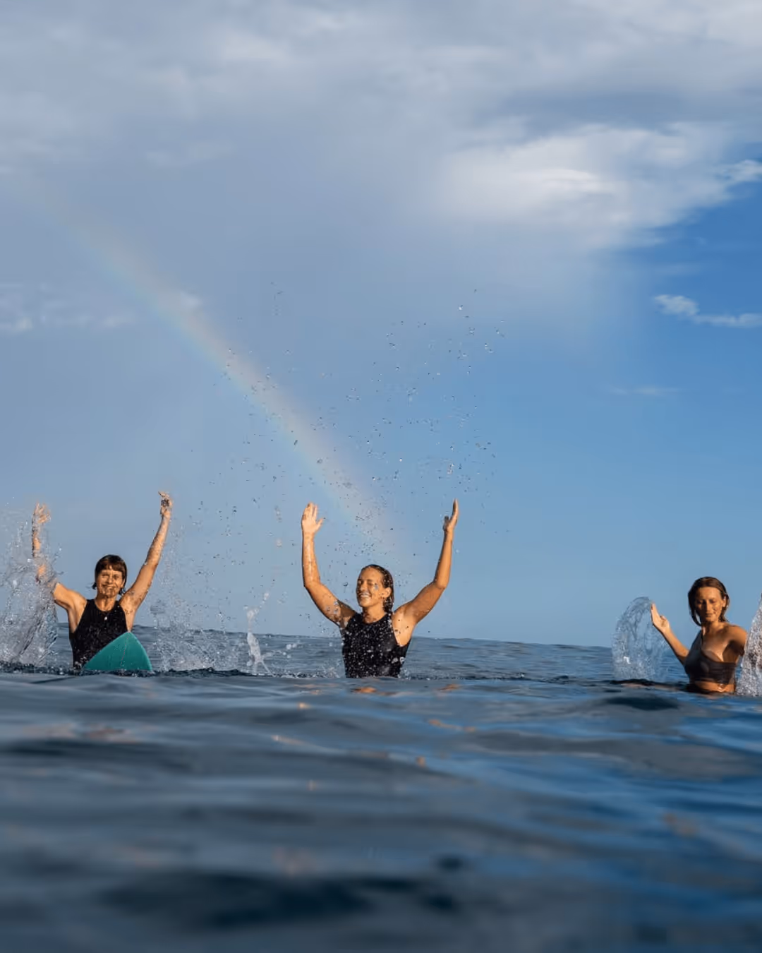 Three people in water raising arms and splashing, with a rainbow in the cloudy blue sky.