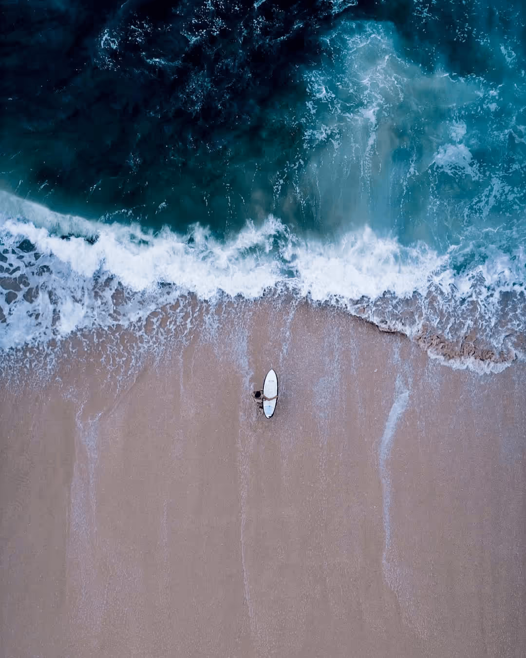 A lone surfer with a white surfboard standing on wet sandy beach near foamy waves.