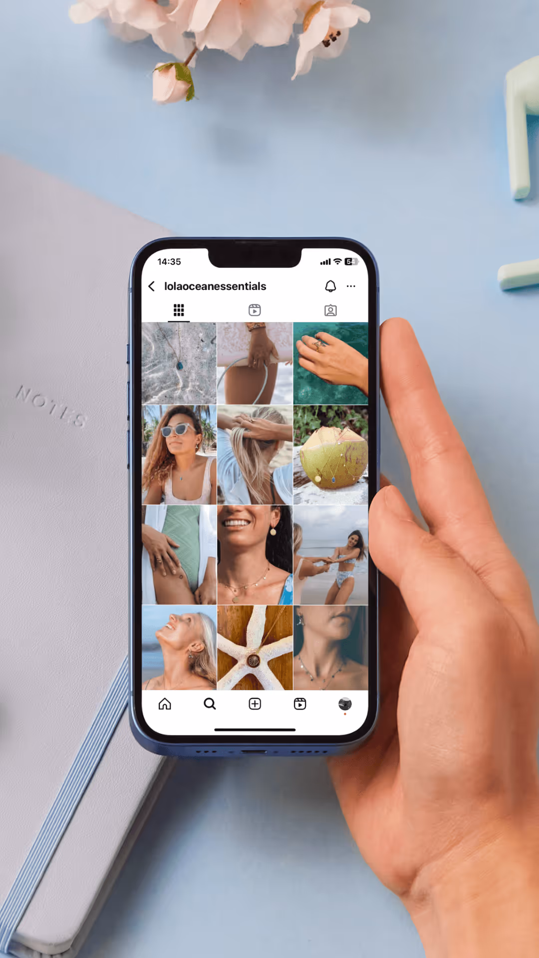 Hand holding phone showing Instagram profile 'lolaoceanessentials' with jewelry and beach photos.