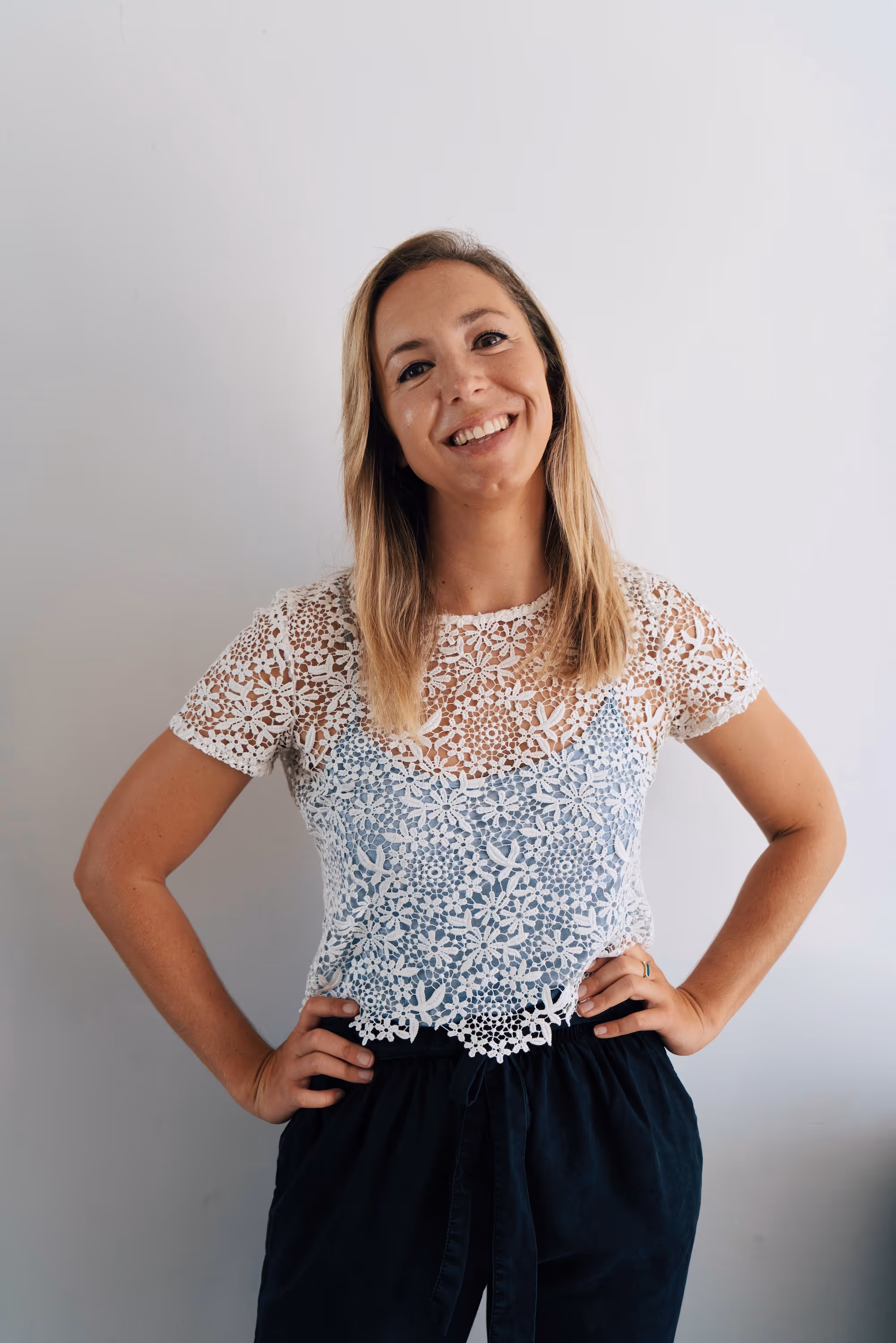 Smiling woman with blonde hair in a white lace top and black pants poses with hands on hips.