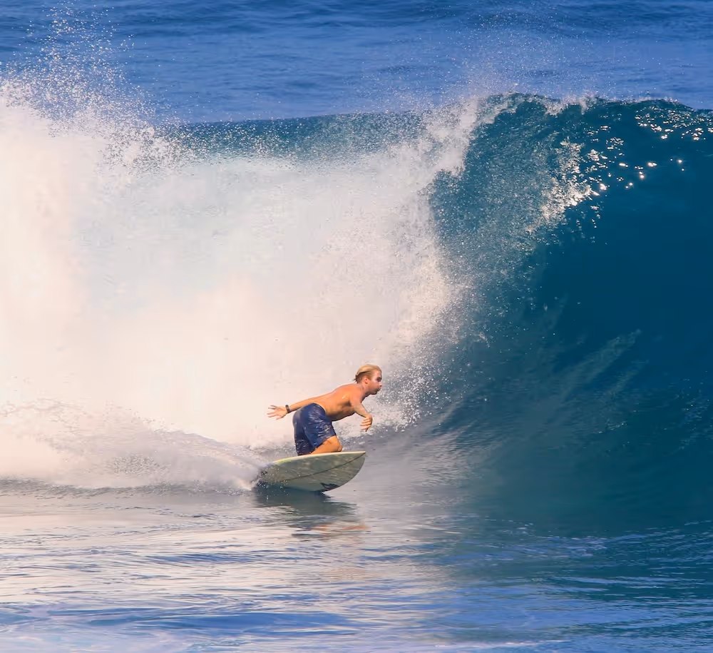 Surfer in blue shorts riding a large wave with white spray in the ocean under clear sky.