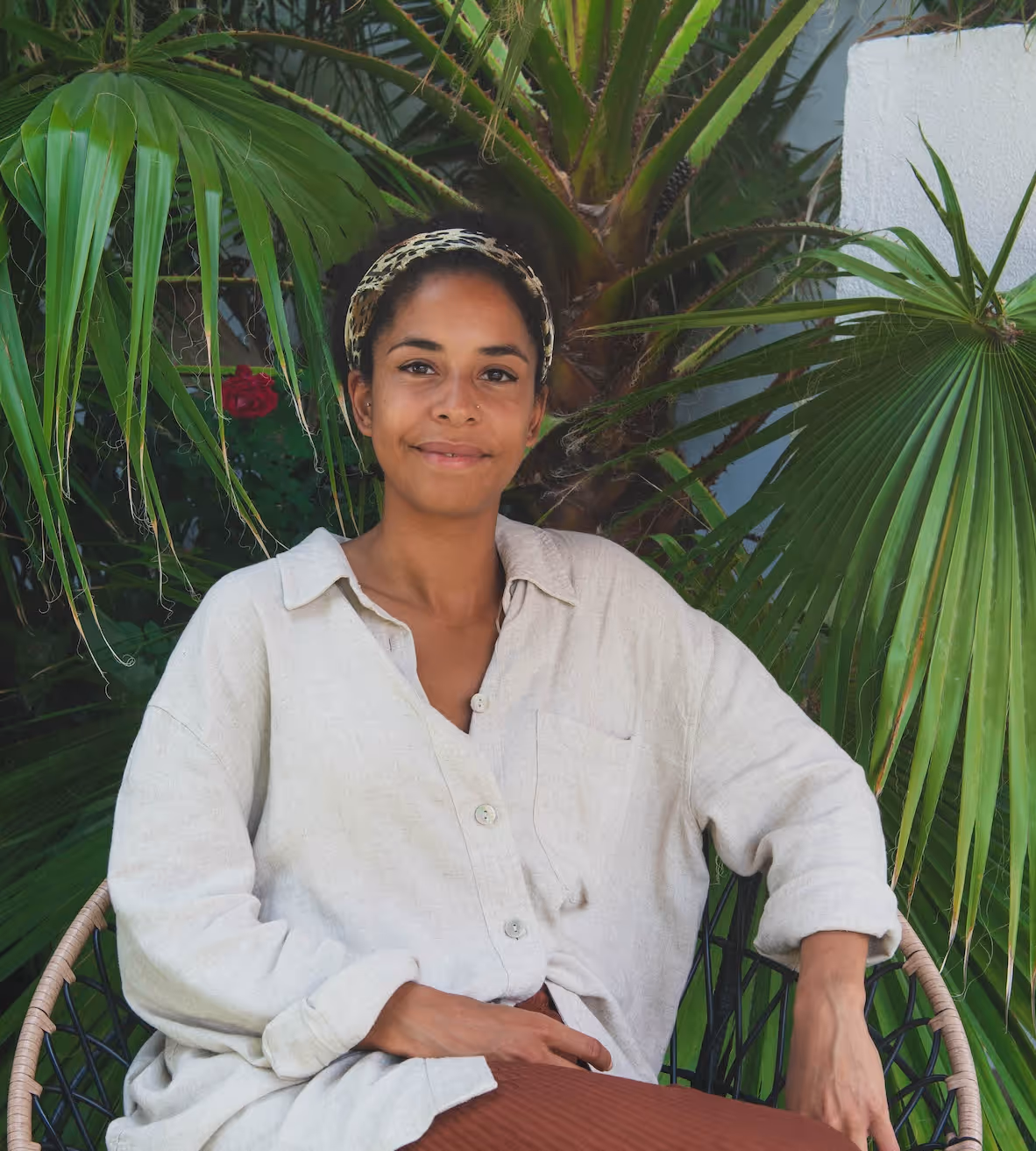Smiling woman wearing a beige shirt and headband seated outdoors near green palm leaves.