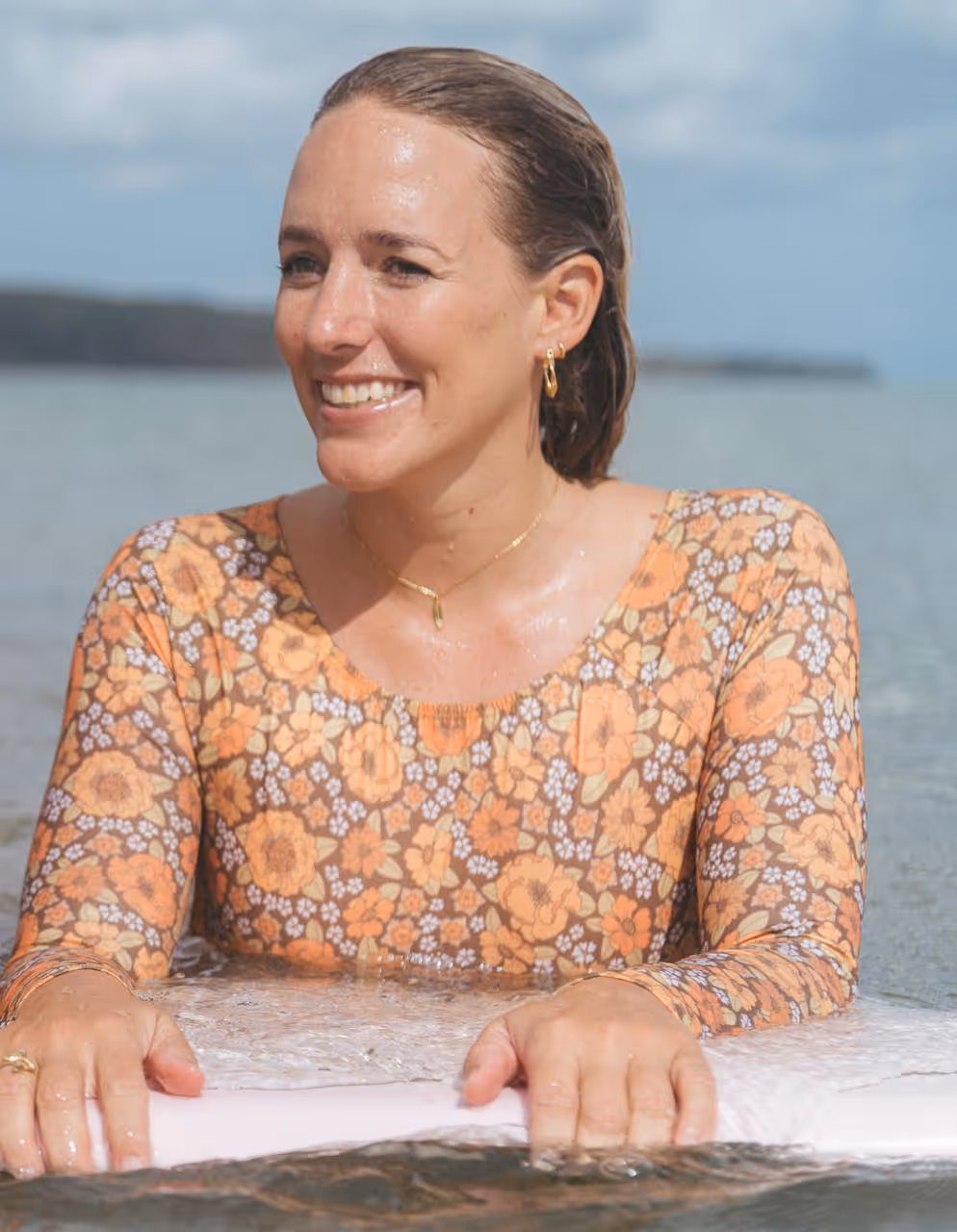 Smiling woman in a floral long-sleeve top holding a surfboard in shallow water.