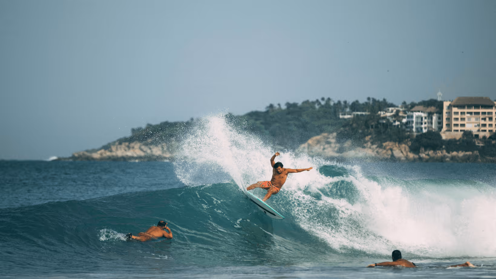 Surfer rides a wave near two others paddling in the ocean with a coastline and buildings in the background.