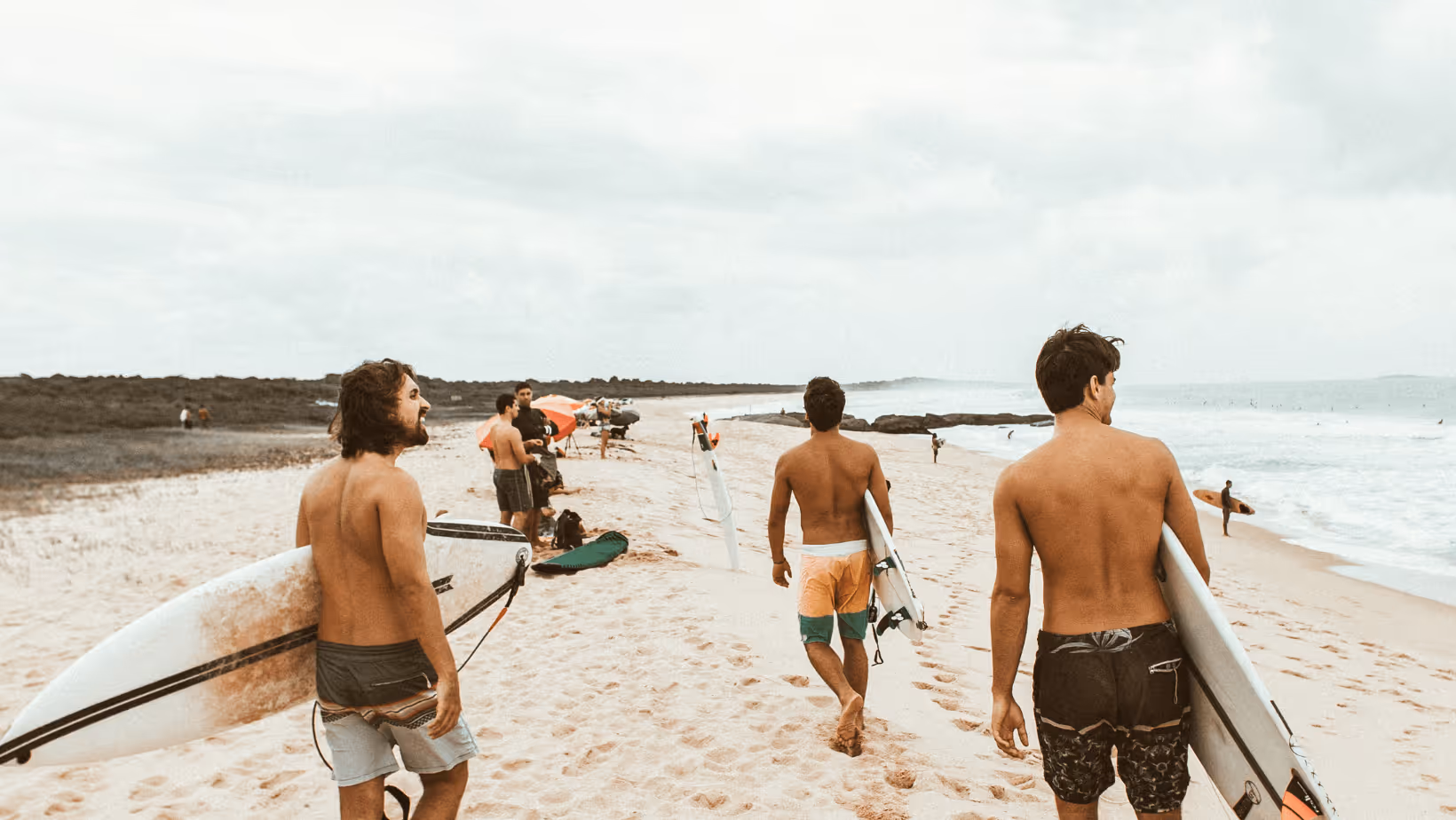 Three shirtless men carrying surfboards walking on a sandy beach toward the ocean.