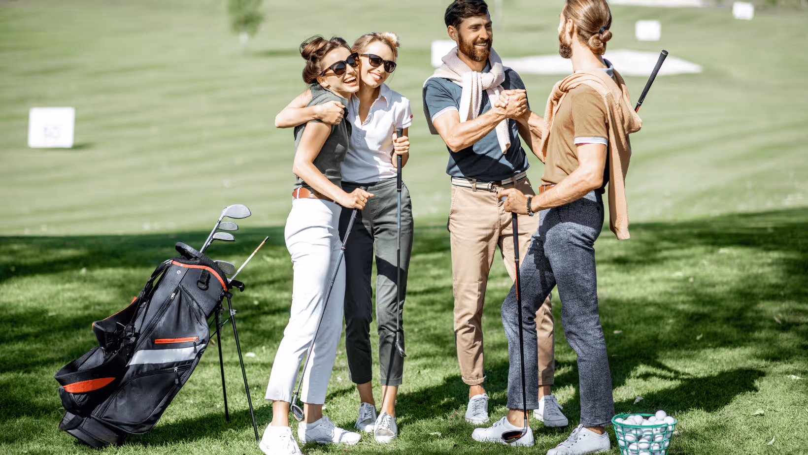 Two women hugging and two men shaking hands on a sunny golf course with clubs and balls nearby.