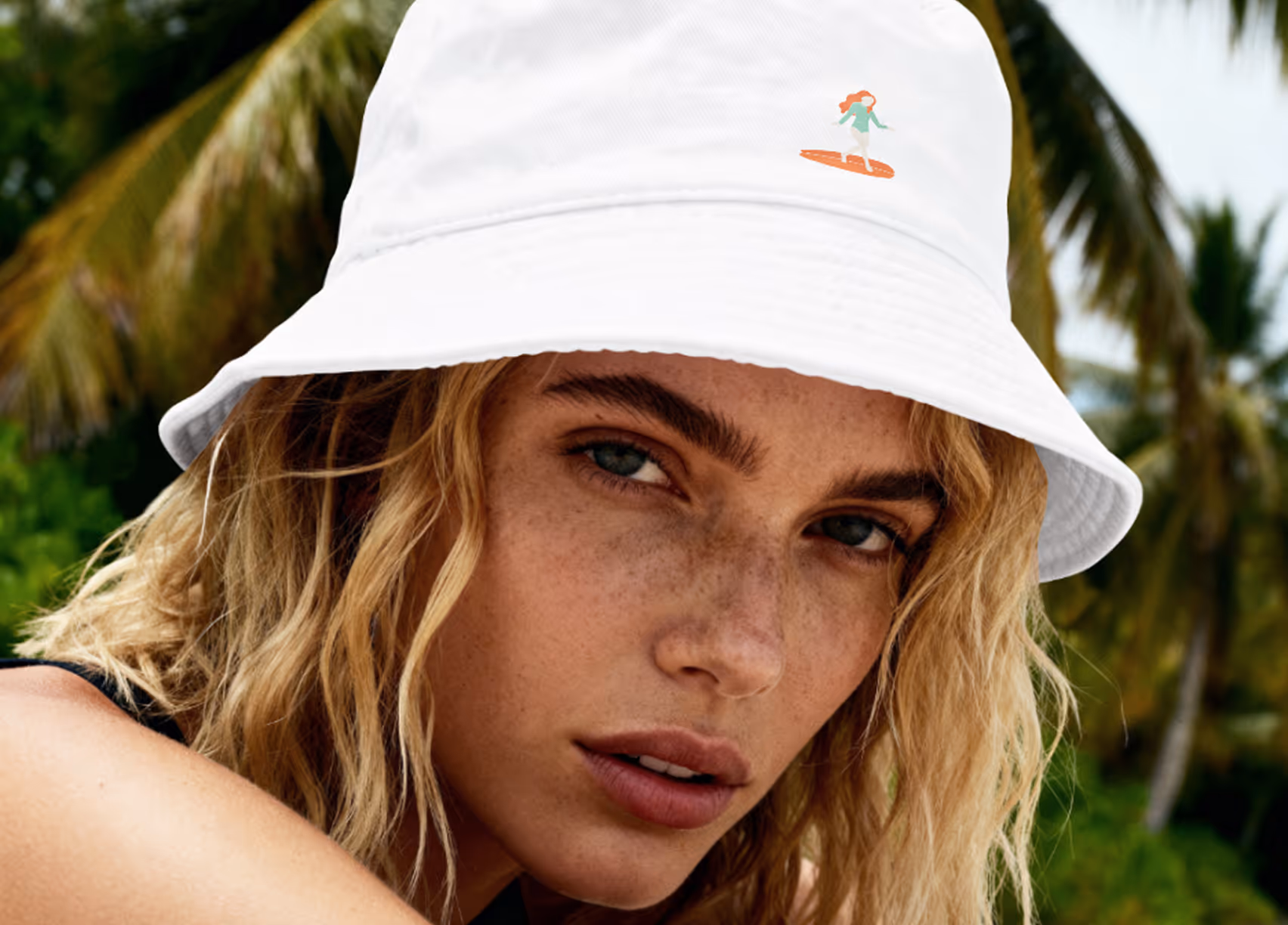 Close-up of a woman with freckles wearing a white bucket hat with a small surfer graphic outdoors.
