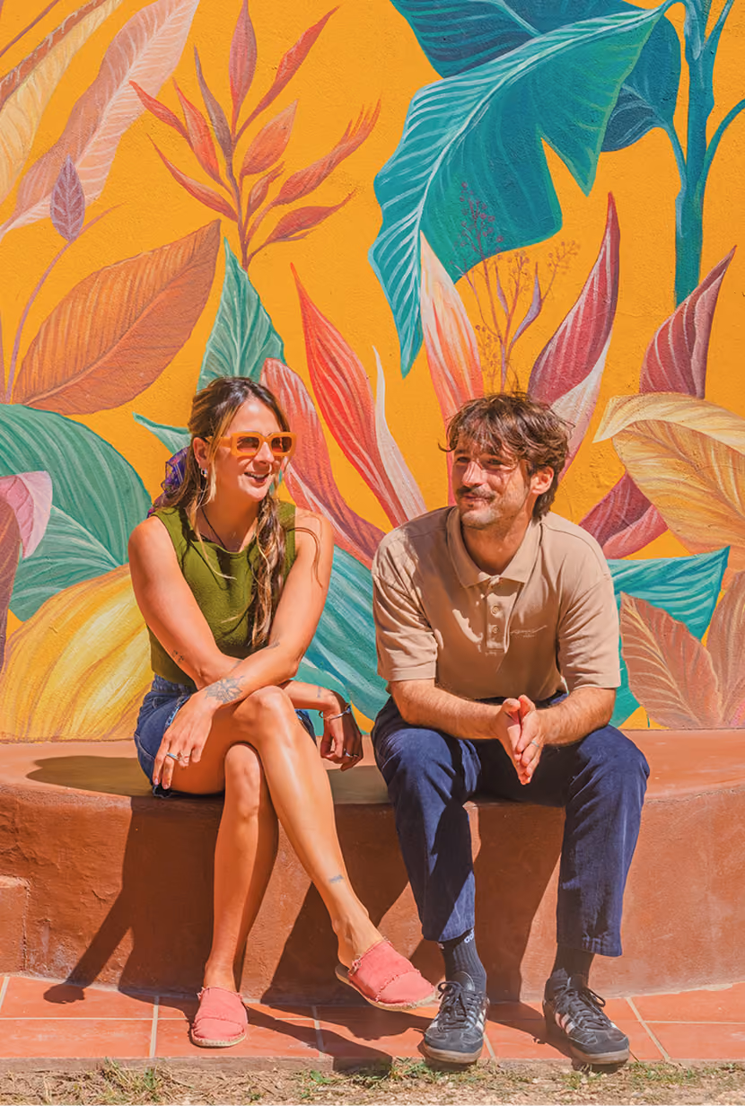 Young woman and man sitting and chatting on a curved bench in front of a colorful mural of large leaves.