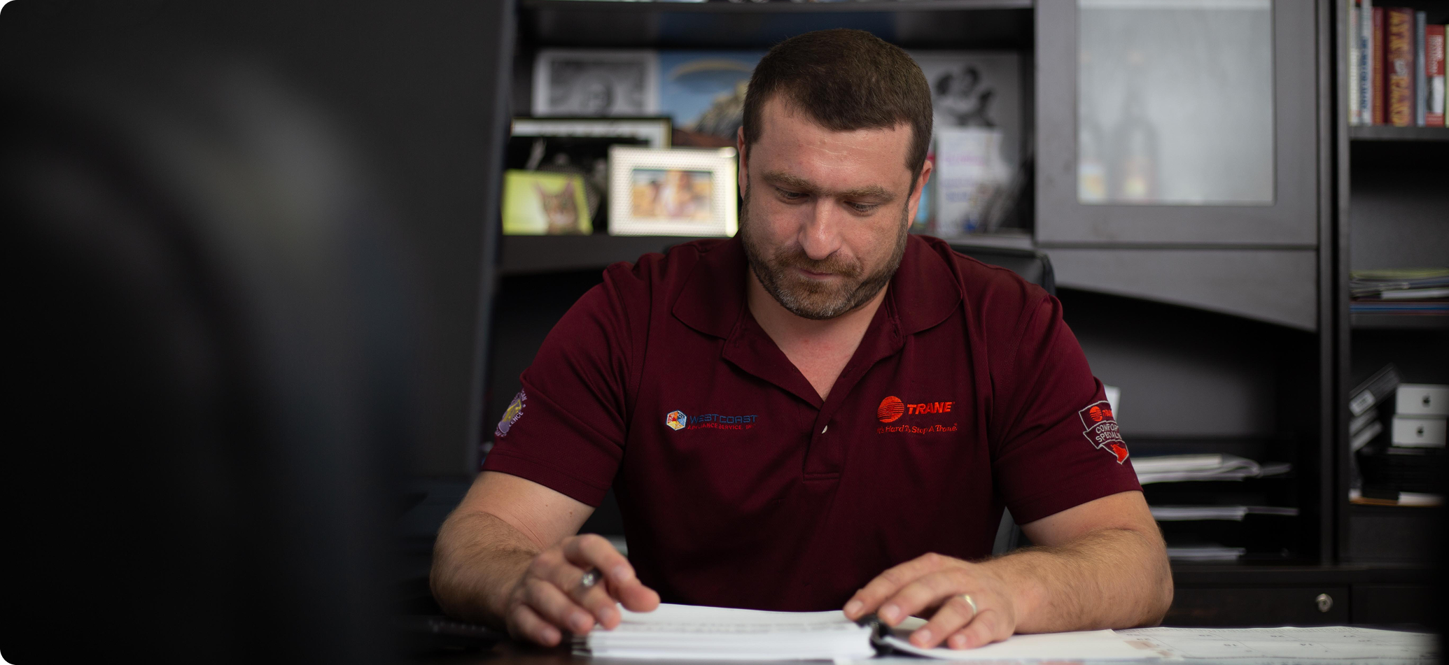 Man in a maroon polo shirt reading and reviewing papers at a desk in an office setting.