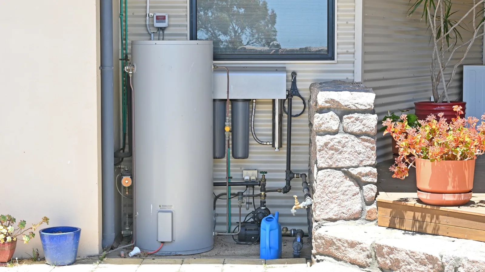 Outdoor water filtration and heating system installed against a corrugated metal wall, with potted plants nearby.