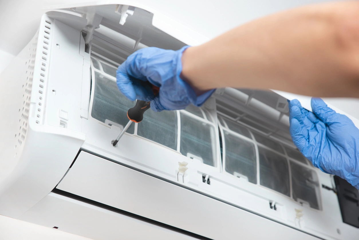 A person wearing blue gloves uses a screwdriver to service an open wall-mounted air conditioner