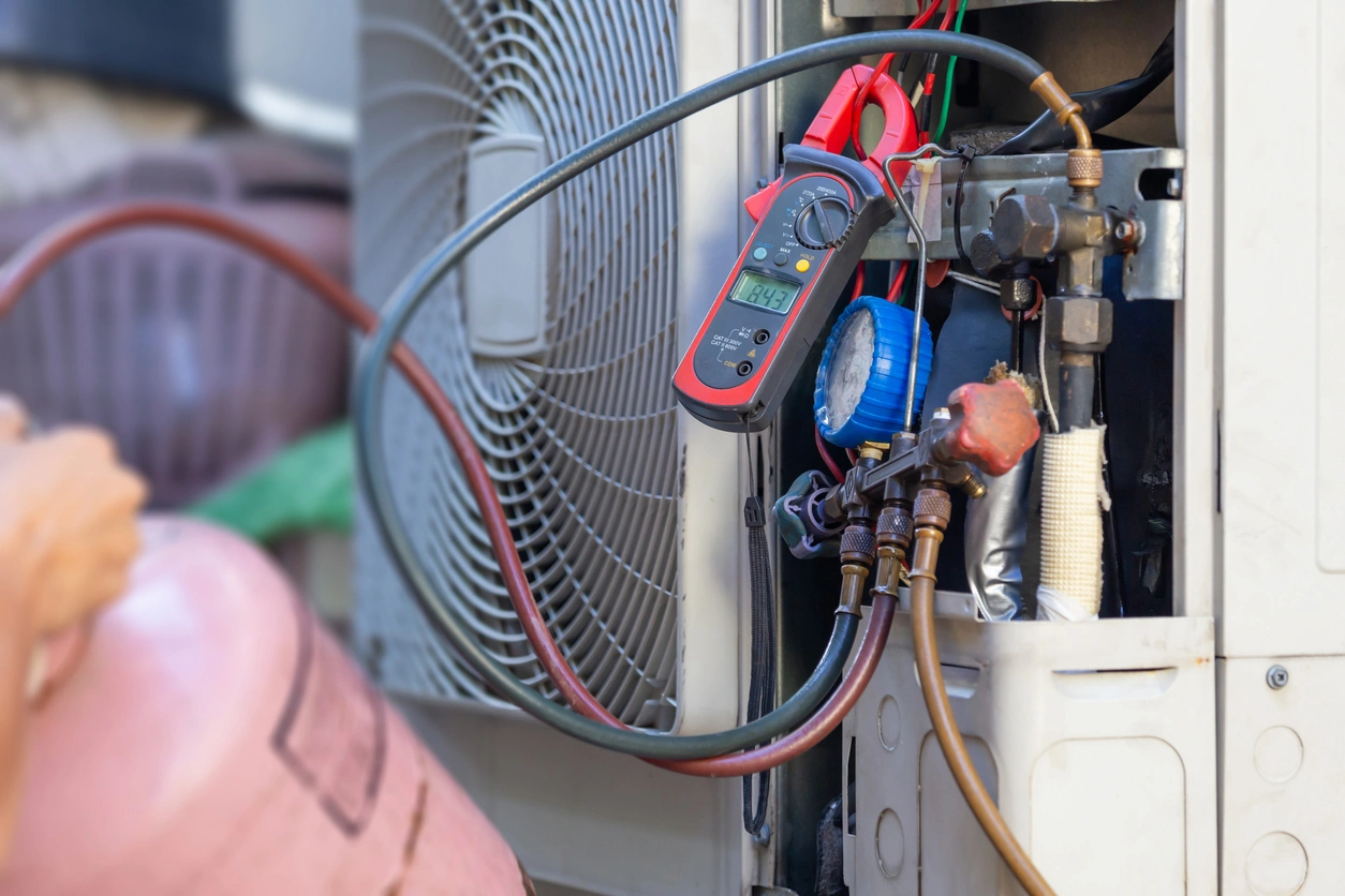 Technician adjusting an air conditioning unit with tools. A red multimeter displays readings, and hoses are connected. 