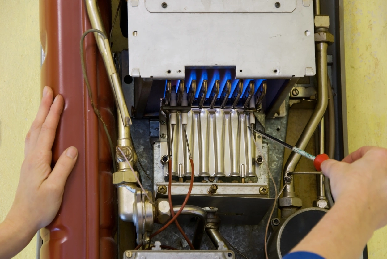 A person in blue sleeves uses a screwdriver to adjust a gas boiler with visible blue flames.