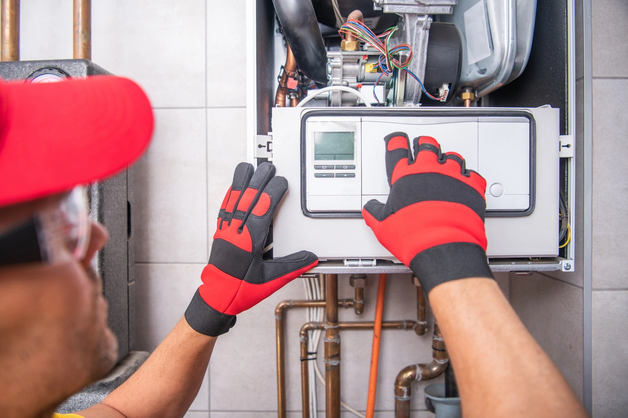 A technician wearing red gloves and hat adjusts a control panel on a furnace. 