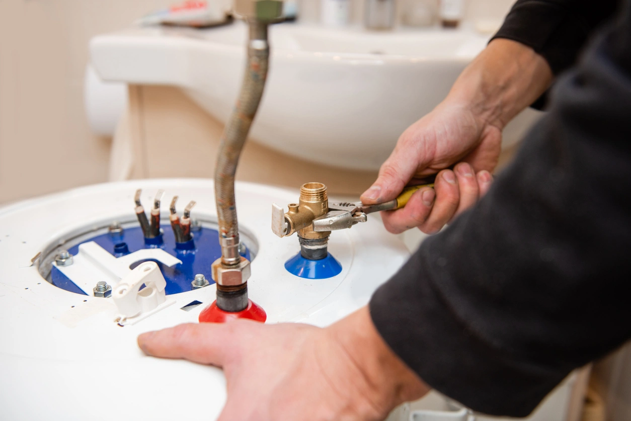Person using a wrench to adjust pipes on an electric water heater, with a sink in the background. 