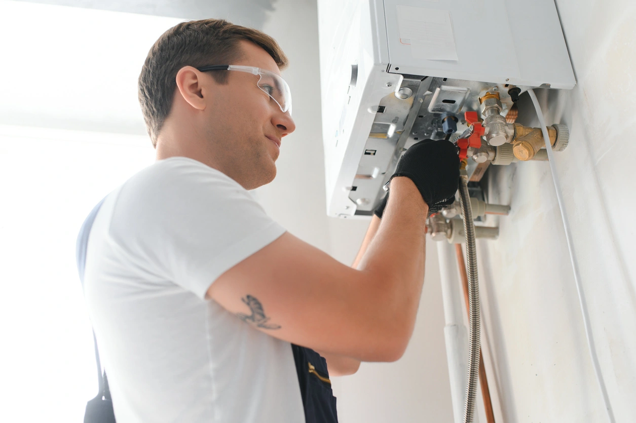 A technician in safety glasses and gloves works on a wall-mounted boiler.