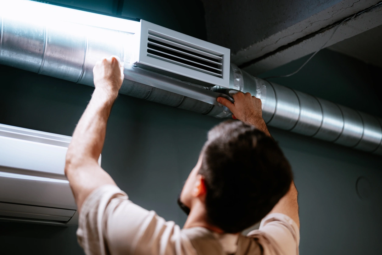 A person adjusts a ceiling air vent connected to a silver duct in a dimly lit room. 