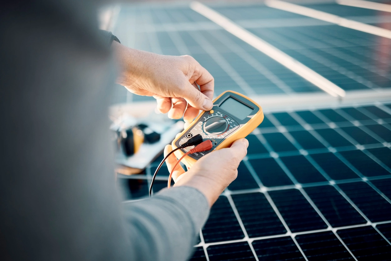 Hands hold a yellow multimeter with red and black probes, testing solar panels.