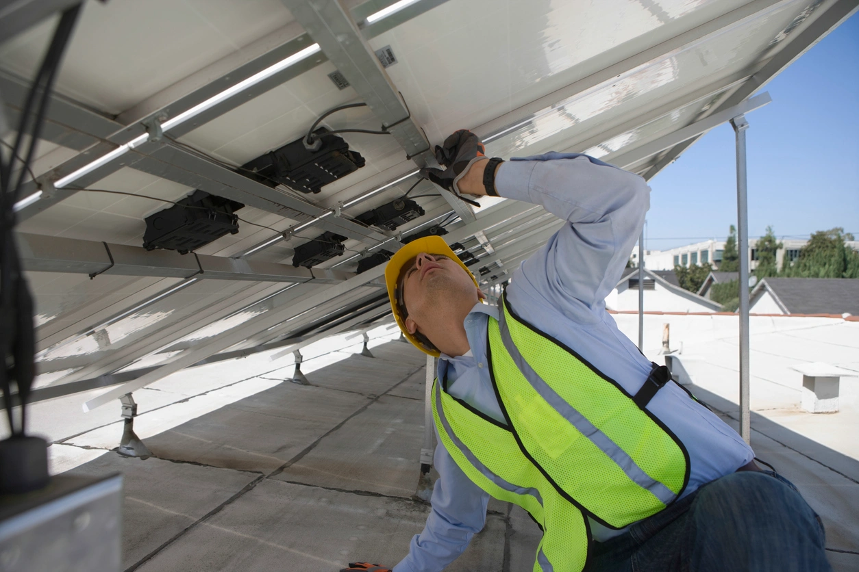 A worker in a yellow hard hat and safety vest inspects a rooftop solar panel.