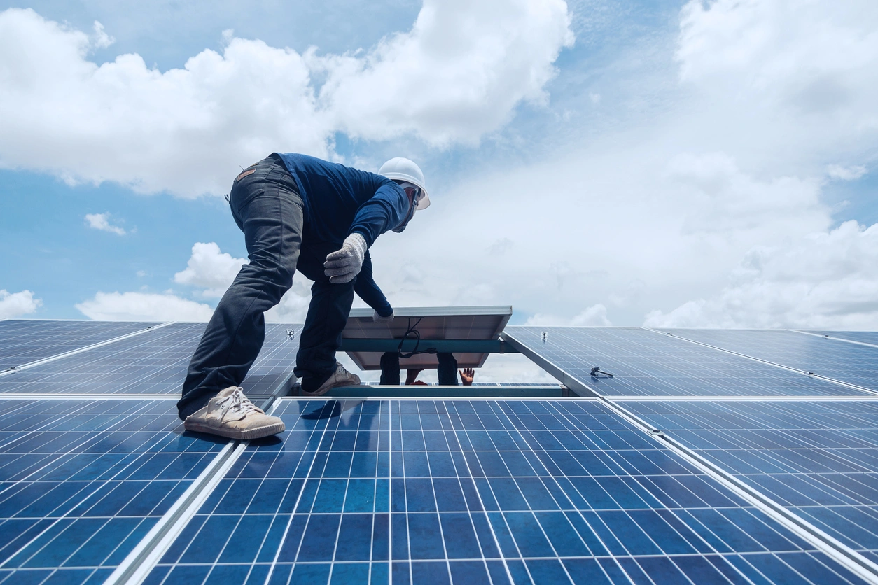 A worker in safety gear adjusts a solar panel on a roof under a bright blue sky with scattered clouds,