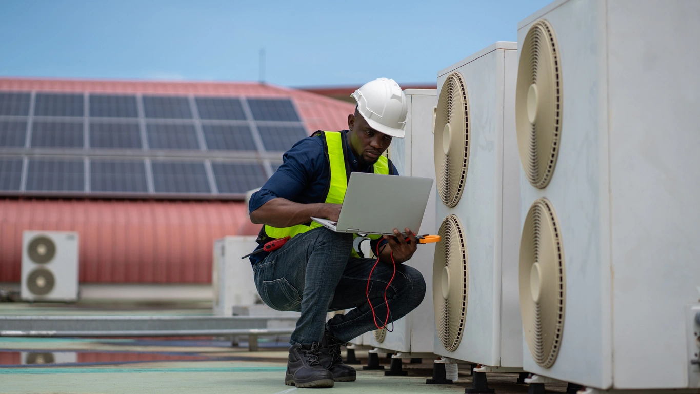 A technician in a white helmet and safety vest kneels on a roof beside air conditioning units, using a laptop. Solar panels are visible in the background.
