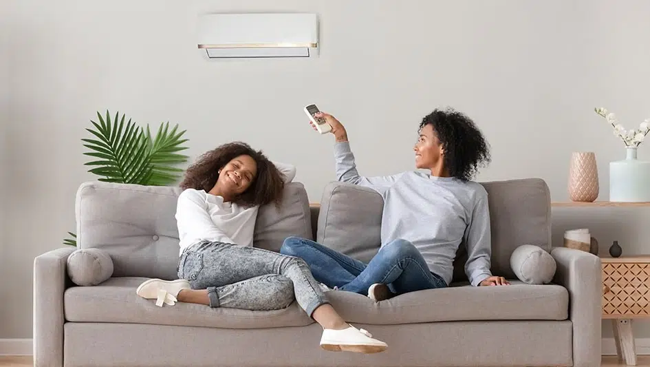Two women relaxing on a gray sofa, one using a remote control, with a ductless mini split air conditioning unit mounted on the wall, emphasizing comfort and efficient climate control in a modern living space.