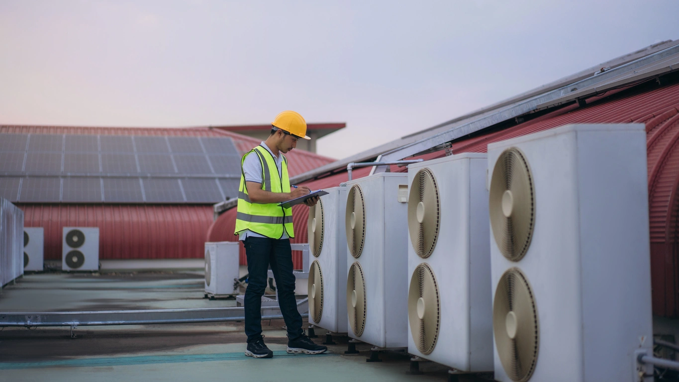 A worker in a yellow vest and hard hat inspects rooftop air conditioning units with large fans, holding a clipboard against a backdrop of solar panels.
