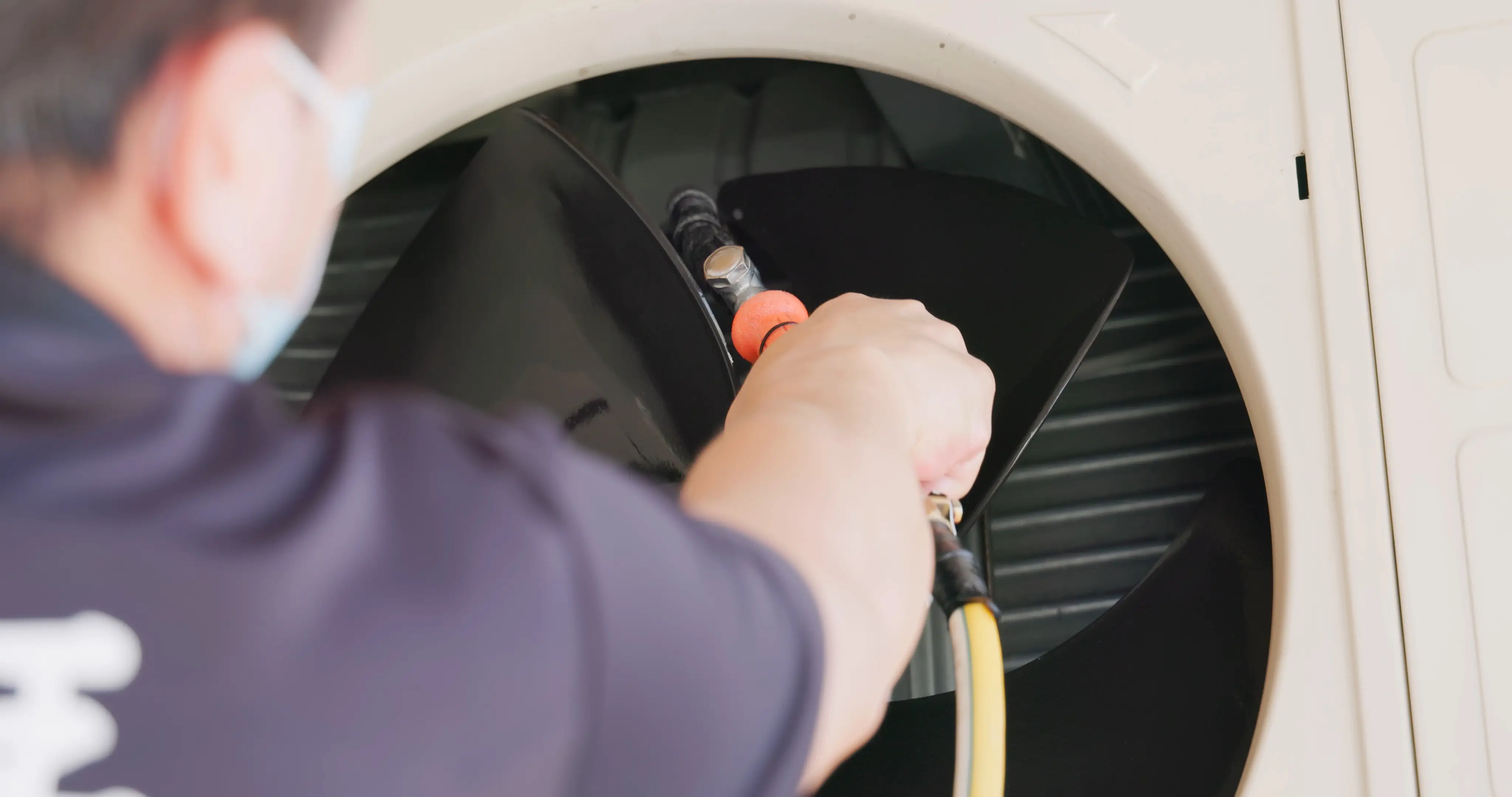 A white tower-style air purifier stands on a polished wooden floor in a home hallway. A small potted plant on a stand is positioned behind it, with an open doorway visible in the background.