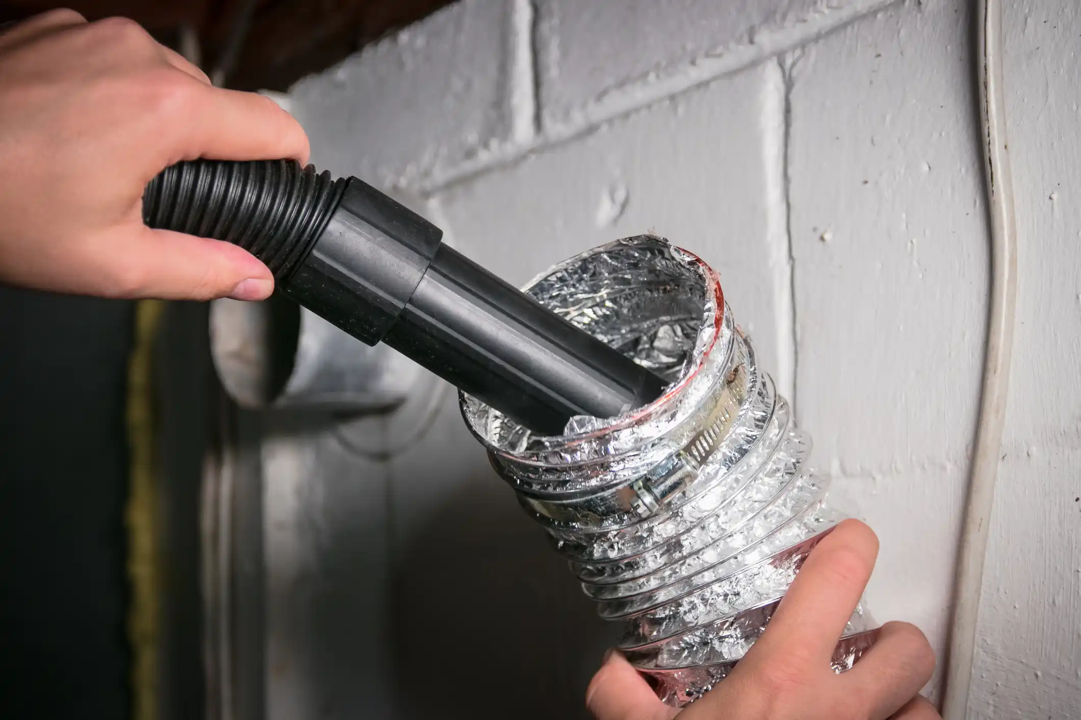 A person's hands insert a black vacuum attachment into a flexible silver dryer vent hose to remove lint. The hose is secured to a white masonry wall with a metal clamp in a basement setting.