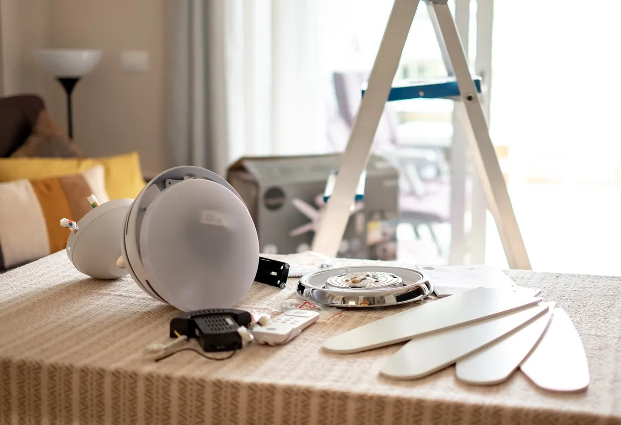 Parts of a white ceiling fan, including the motor housing and blades, lie on a textured tablecloth. A stepladder and fan packaging are visible in the blurred background of a brightly lit living room.