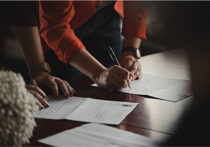 A person in a red blazer signs important documents on a wooden table.