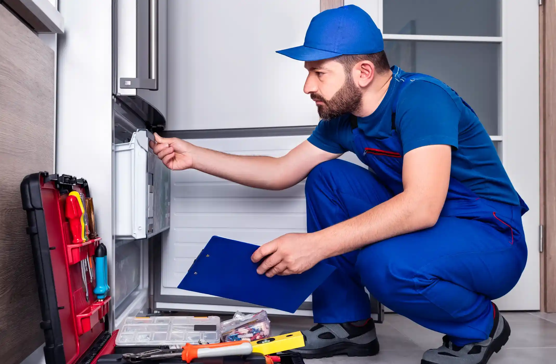 A male technician wearing a blue uniform and cap crouches with a clipboard and toolbox, repairing a commercial refrigerator drawer.