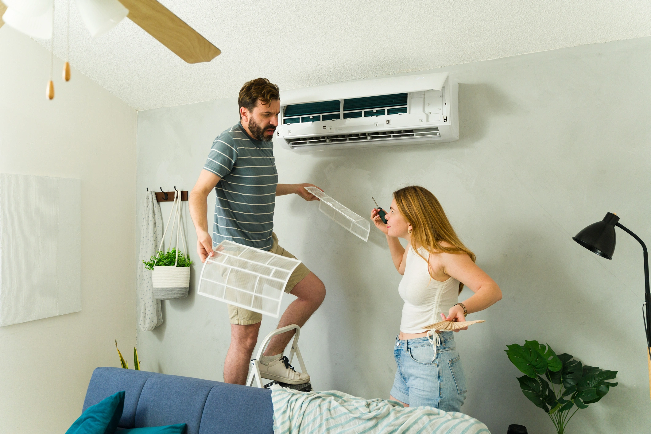 A man on a ladder holds air conditioner filters while a woman hands him a screwdriver. 