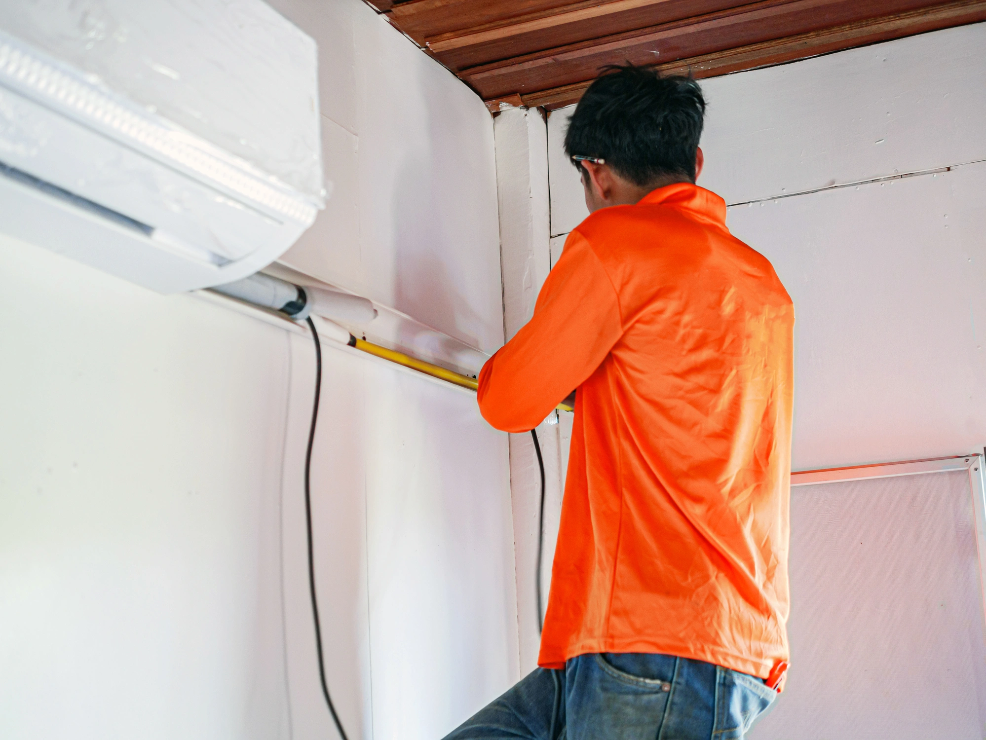 A person in an orange shirt is using a yellow tape measure on a white wall near an air conditioning unit