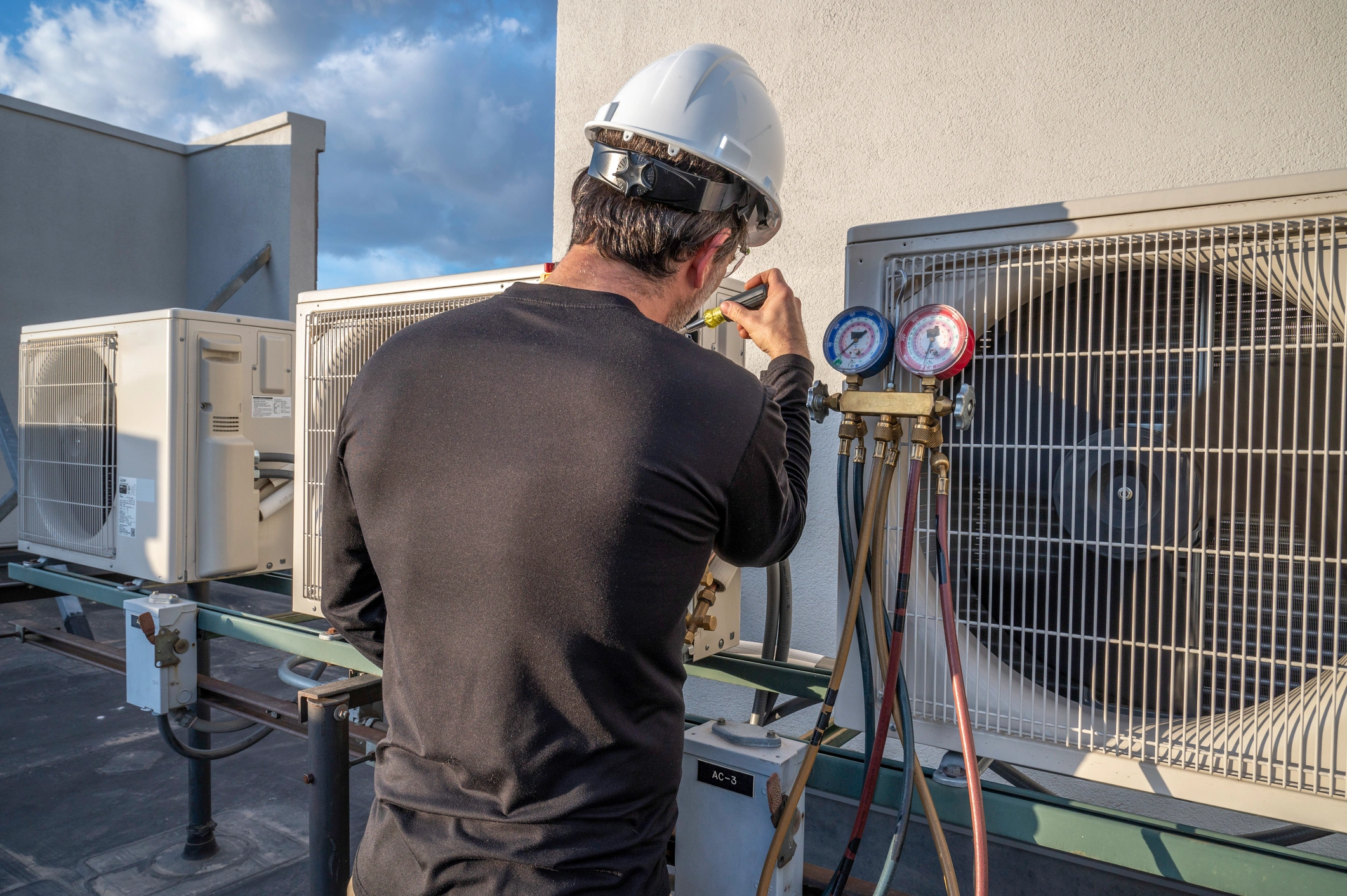 A technician in a hard hat and black shirt works on rooftop air conditioning units