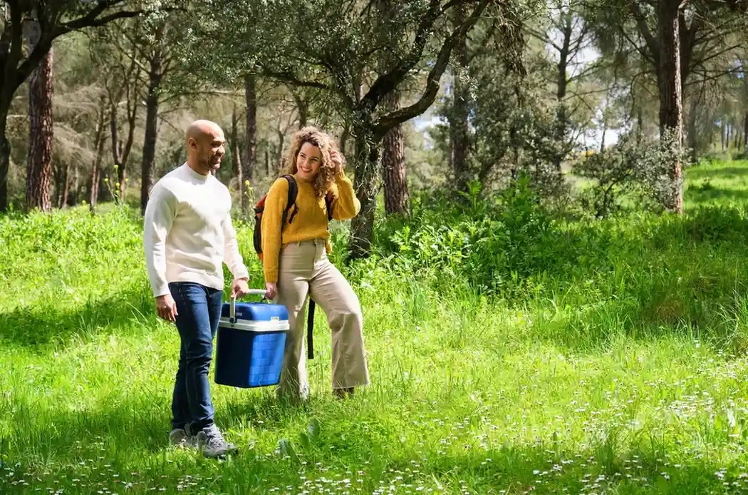A smiling couple walk through a grassy, sunlit forest area; the man carries a blue cooler, and the woman wears a backpack.