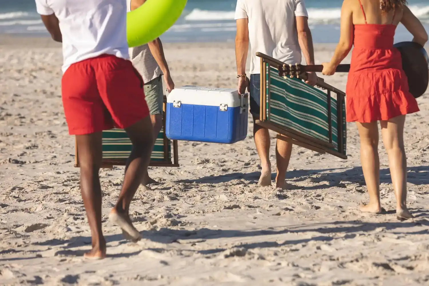 People walk across a sandy beach toward the ocean, carrying a blue cooler, folding beach chairs, and an inflatable ring.