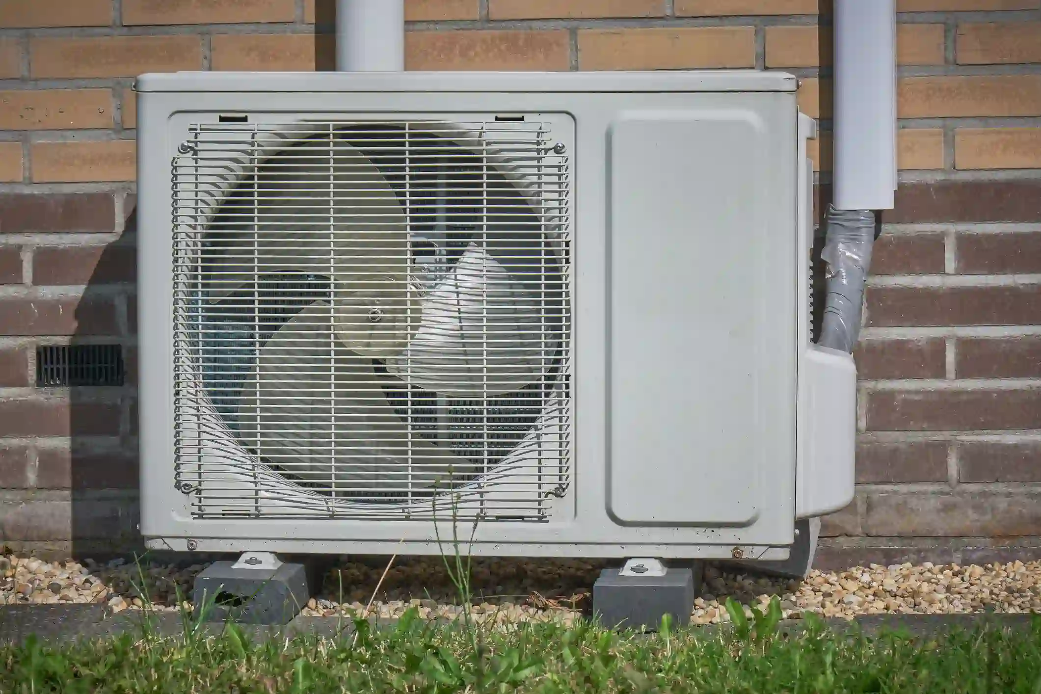 A white, outdoor mini-split heat pump or air conditioning unit is mounted on gray blocks and sits in front of a red brick wall. The unit's large fan is visible behind a protective grille. White plastic conduit runs down the wall on the right side, connecting to the unit. The foreground shows a strip of green grass and a gravel bed.