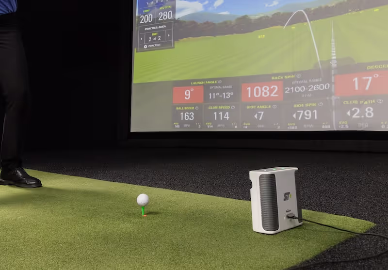 Golf ball teed on an indoor mat with a SKYTRAK plus launch monitor, aimed toward GOLFTEC Bay simulation studio 