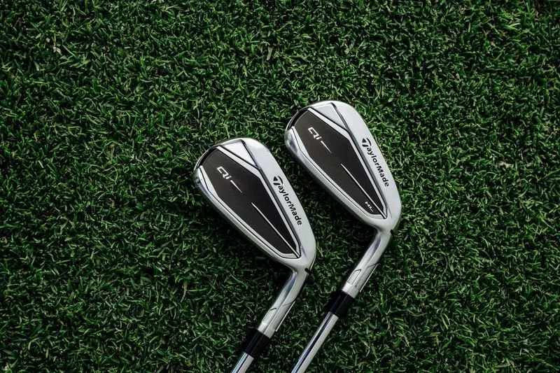 Close-up view of two TaylorMade Qi iron clubheads on a grassy background.