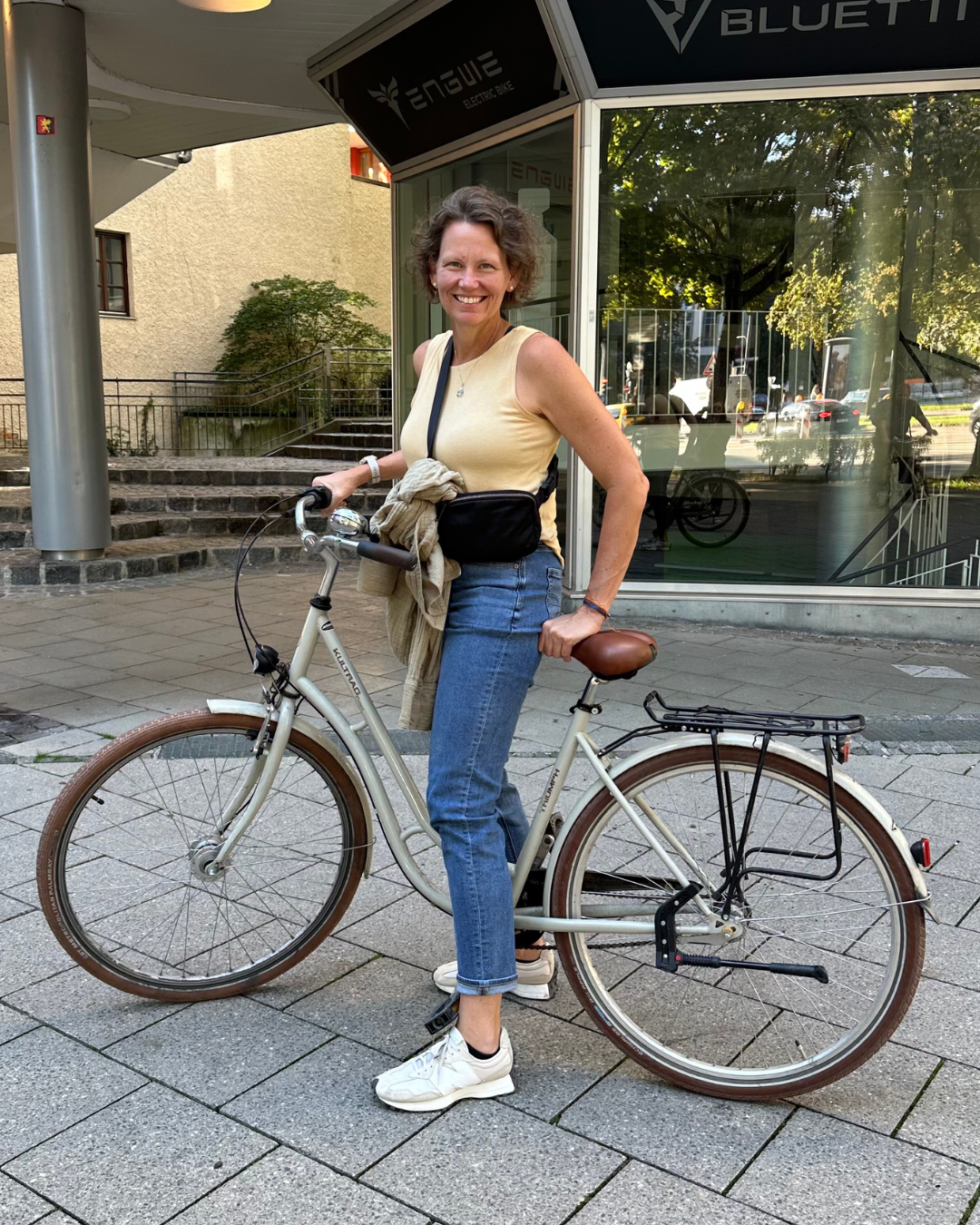Laura Fletcher   wearing a yellow tank top and jeans stands outdoors with her bicycle on a paved sidewalk. She has a light sweater tied around her waist and a small black bag across her chest, with buildings and trees visible in the background.