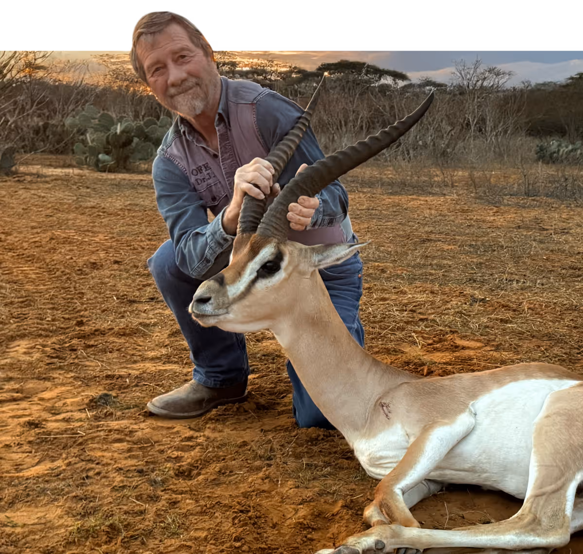 Smiling man kneels beside antelope with curved horns in dry landscape