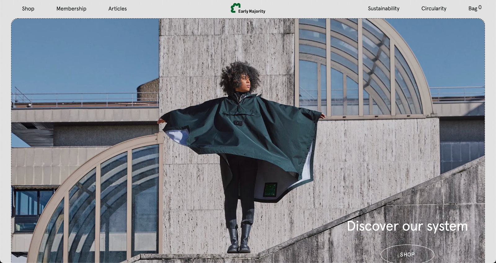 The Early Majority homepage, featuring a Black woman modeling a green poncho, black pants, and black calf-height boots with her arms stretched out. She is centered on the page and standing in front of a concrete building with brutalist architectural style. The bottom right of the page features a "Shop" CTA button in an oval below the text, "Discover our system."