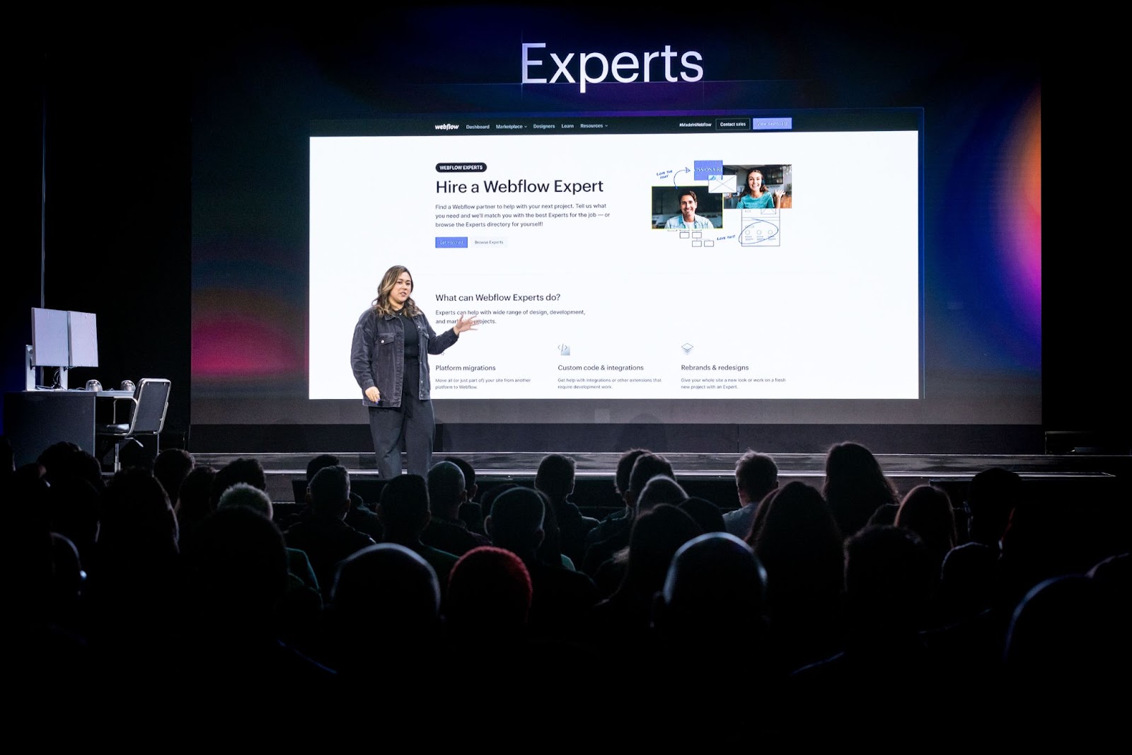 Emily Lonetto presenting on stage in front of a live crowd at Webflow Conf 22. The presentation screen behind her displays the Webflow Experts website page.