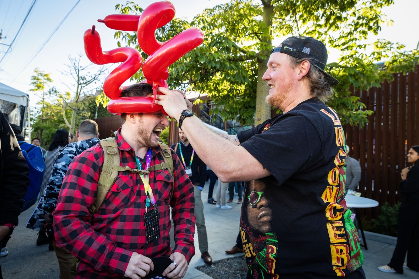 A man with a backwards baseball hat puts a balloon hat on another man.